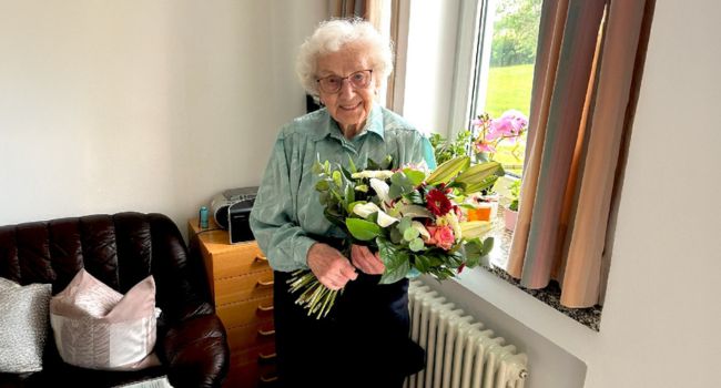 Ältere Frau hält bunten Blumenstrauß in hellem Wohnzimmer mit Sofa und Fenster. Feierliche Stimmung​​​​​​​​​​​​​​​​​​​​​​​​​​​​​​​​​​​​​​​​​​​​​​