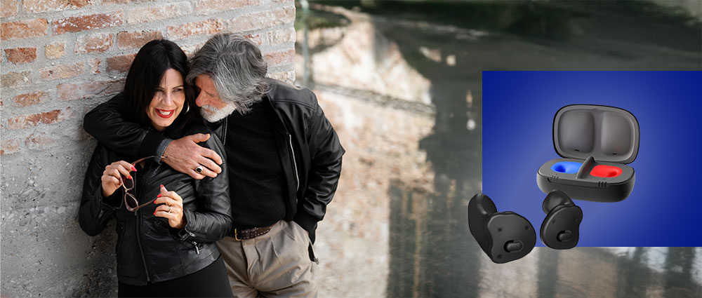 Älteres Paar in Lederjacken lehnt an Backsteinwand, daneben kabellose In-Ear-Kopfhörer im Ladecase vor blauem Hintergrund​​​​​​​​​​​​​​​​​​​​​​​​