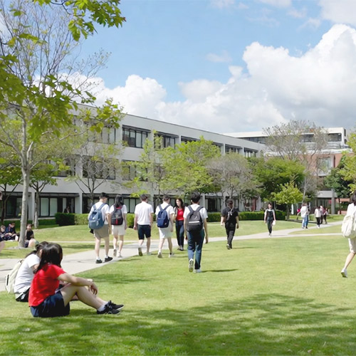 Mehrere Studenten mit Rucksäcken gehen und sitzen auf einer grünen Wiese vor einem modernen Schulgebäude bei klarem Himmel​​​​​​​​​​​​​​​​​​​​​​​