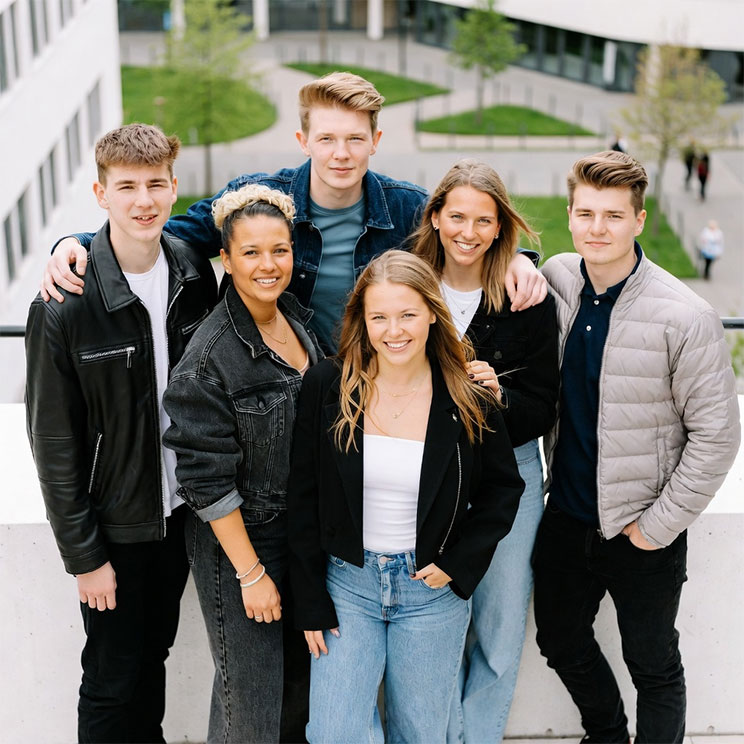 Sechs junge Erwachsene stehen lächelnd vor einem modernen Campus, symbolisieren Ausbildung und Teamgeist.​​​​​​​​​​​​​​​​​​​​​​​​​​​​​​​​​​​​​​​​