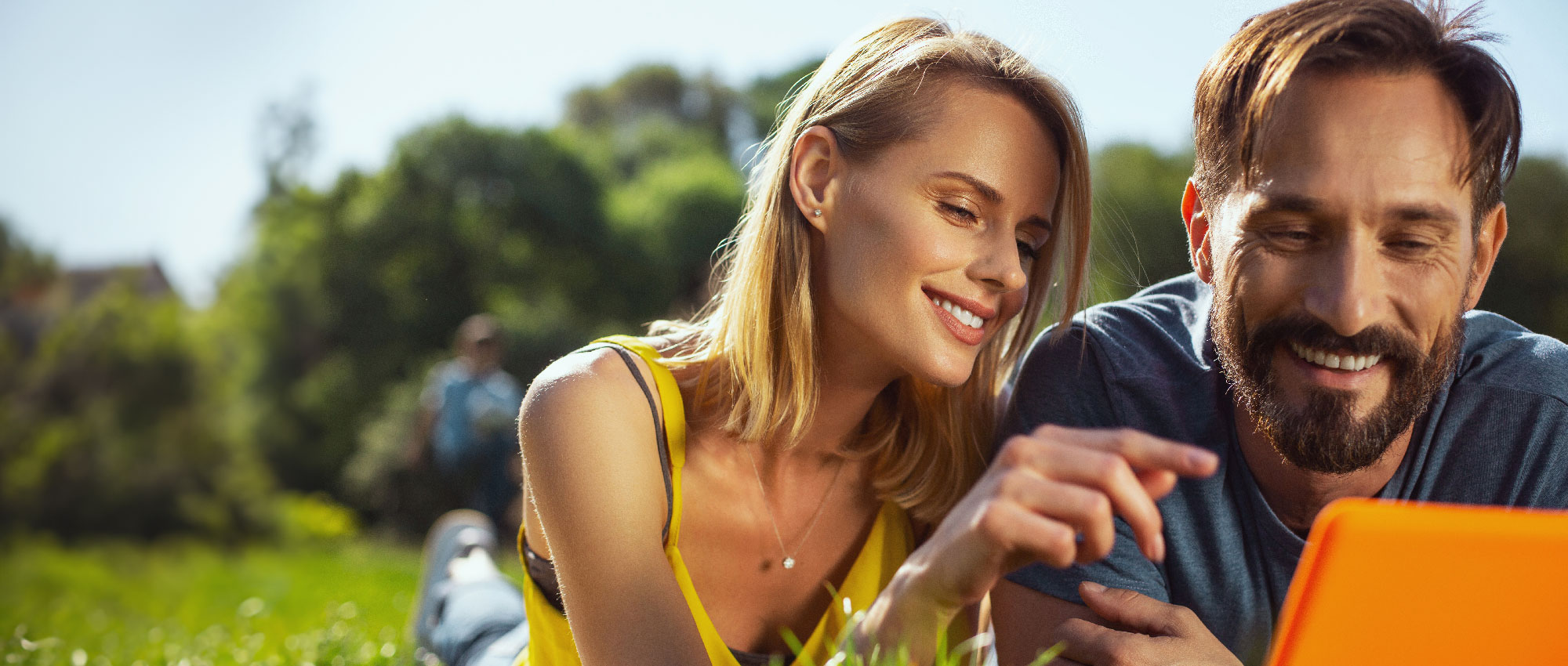 Junges Paar entspannt im Grünen und nutzt ein orangefarbenes Tablet bei sonnigem Wetter im Park​​​​​​​​​​​​​​​​​​​​​​​​​​​​​​​​​​​​​​​​​​​​​​​​​​