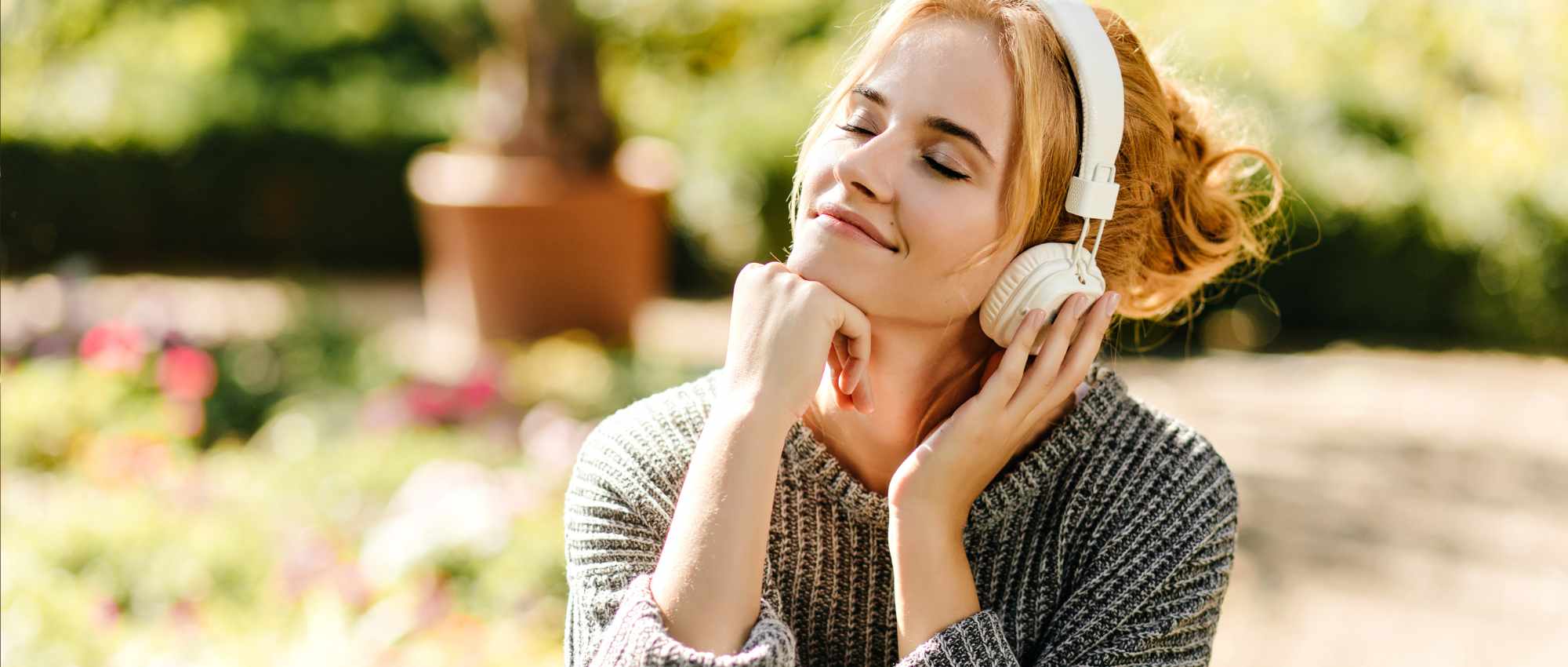 Junge Frau mit geschlossenen Augen hört Musik mit weißen Kopfhörern im sonnigen Garten und wirkt entspannt.​​​​​​​​​​​​​​​​​​​​​​​​​​​​​​​​​​​​​​