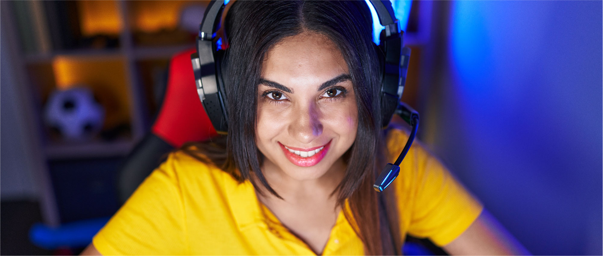 Junge Frau mit gelbem Shirt und Gaming-Headset sitzt vor blau beleuchtetem Hintergrund auf rotem Gaming-Stuhl​​​​​​​​​​​​​​​​​​​​​​​​​​​​​​​​​​​​