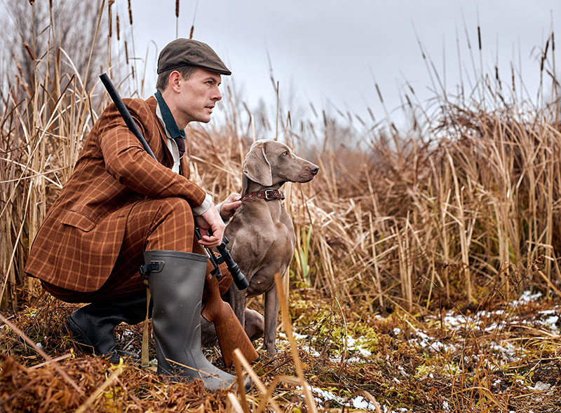 Jäger mit Flinte und Hund im herbstlichen Schilf, umgeben von trockenem Gras und leichtem Schneefall.​​​​​​​​​​​​​​​​​​​​​​​​​​​​​​​​​​​​​​​​​​​​