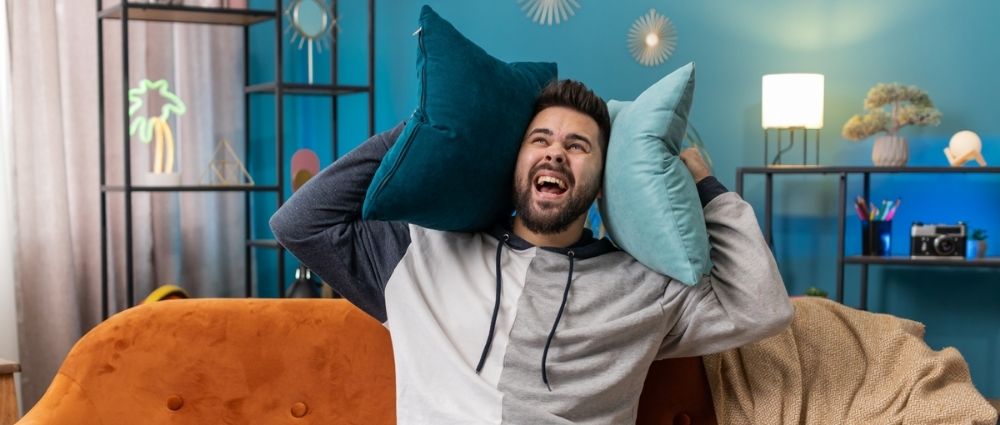 Junger Mann sitzt auf orangefarbenem Sofa und hält zwei Kissen an die Ohren in modern eingerichteter Wohnung mit blauer Wand.​​​​​​​​​​​​​​​​​​​​