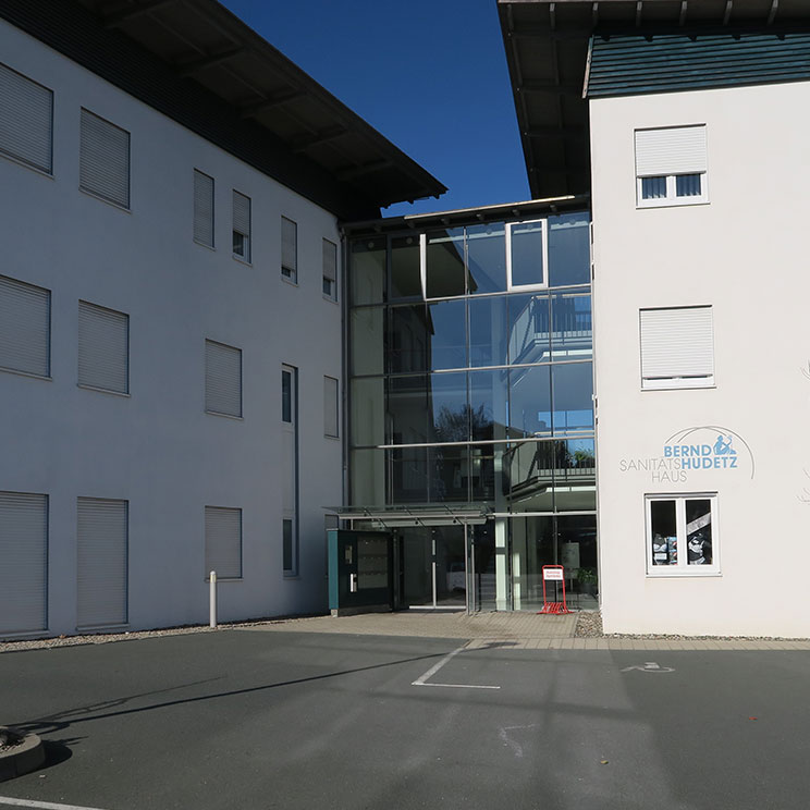 Modernes Sanitätshaus Bernd Hudetz mit großer Glasfassade, Parkplätzen und klarer Architektur bei Sonnenschein​​​​​​​​​​​​​​​​​​​​​​​​​​​​​​​​​​​