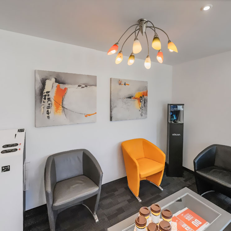 Moderner Wartebereich mit grauen und orangen Sesseln, abstrakten Bildern und stilvoller Beleuchtung im Hörakustiker-Büro.​​​​​​​​​​​​​​​​​​​​​​​​