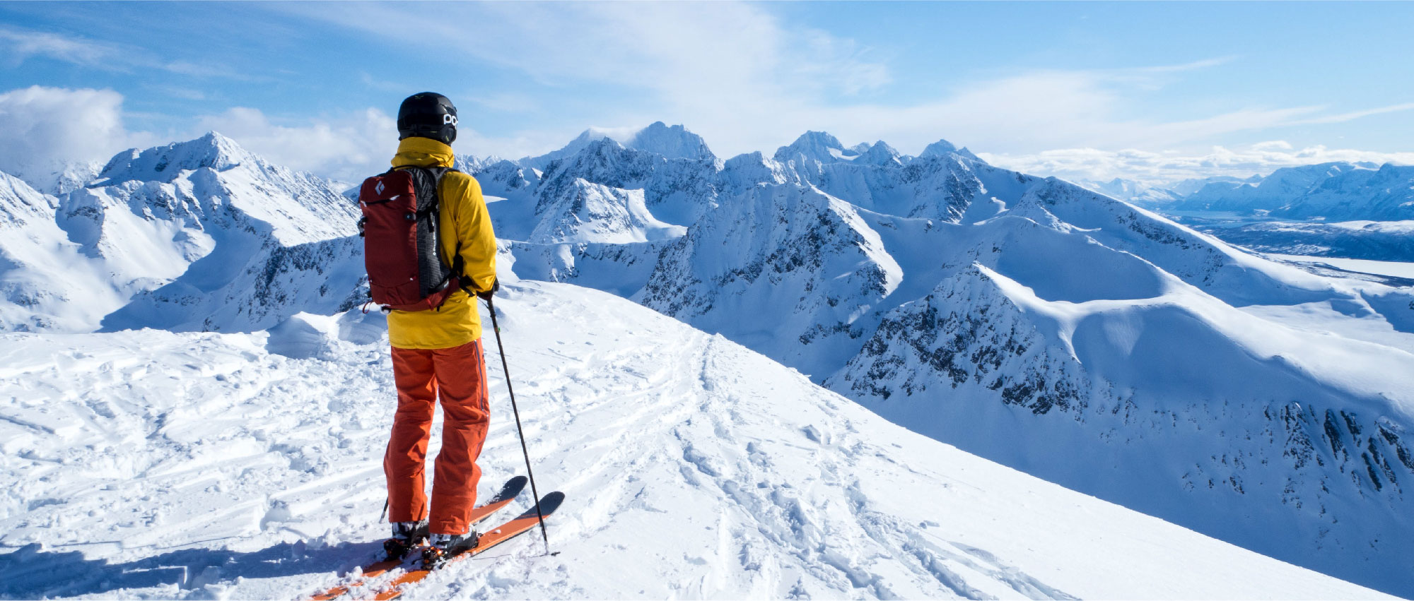 Skifahrer in gelber Jacke und roter Hose blickt auf verschneite Berglandschaft bei klarem Himmel im Wintersportgebiet.​​​​​​​​​​​​​​​​​​​​​​​​​​​