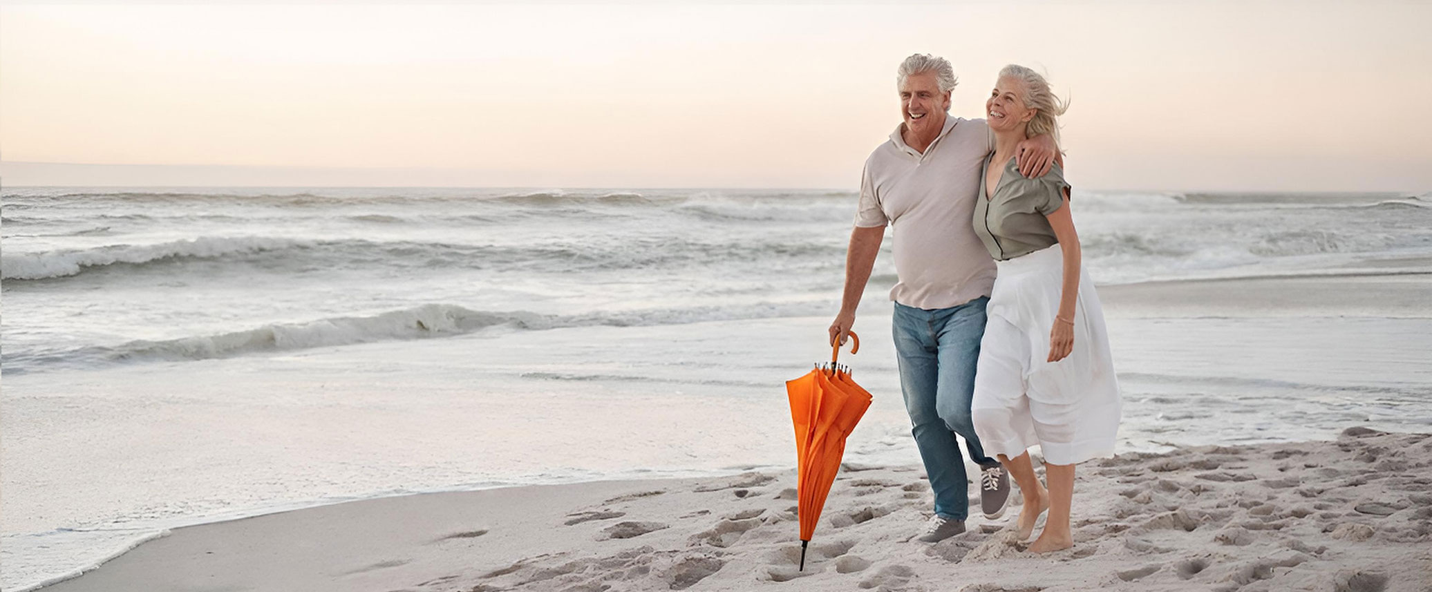 Älteres Paar geht am Strand spazieren, hält orangefarbenen Regenschirm, entspannte Stimmung am Meer​​​​​​​​​​​​​​​​​​​​​​​​​​​​​​​​​​​​​​​​​​​​​​