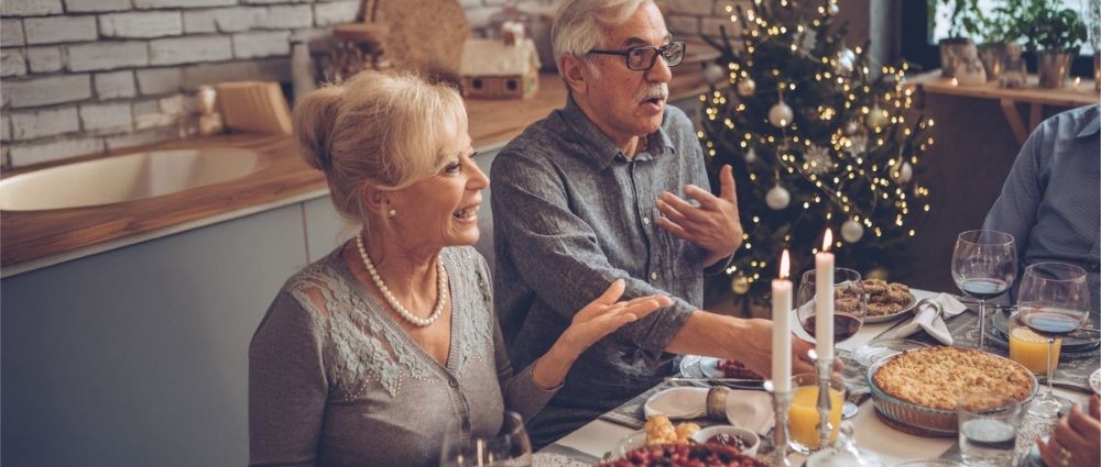 Älteres Paar unterhält sich lebhaft beim festlichen Abendessen mit Kerzenlicht und Weihnachtsbaum im Hintergrund​​​​​​​​​​​​​​​​​​​​​​​​​​​​​​​​​