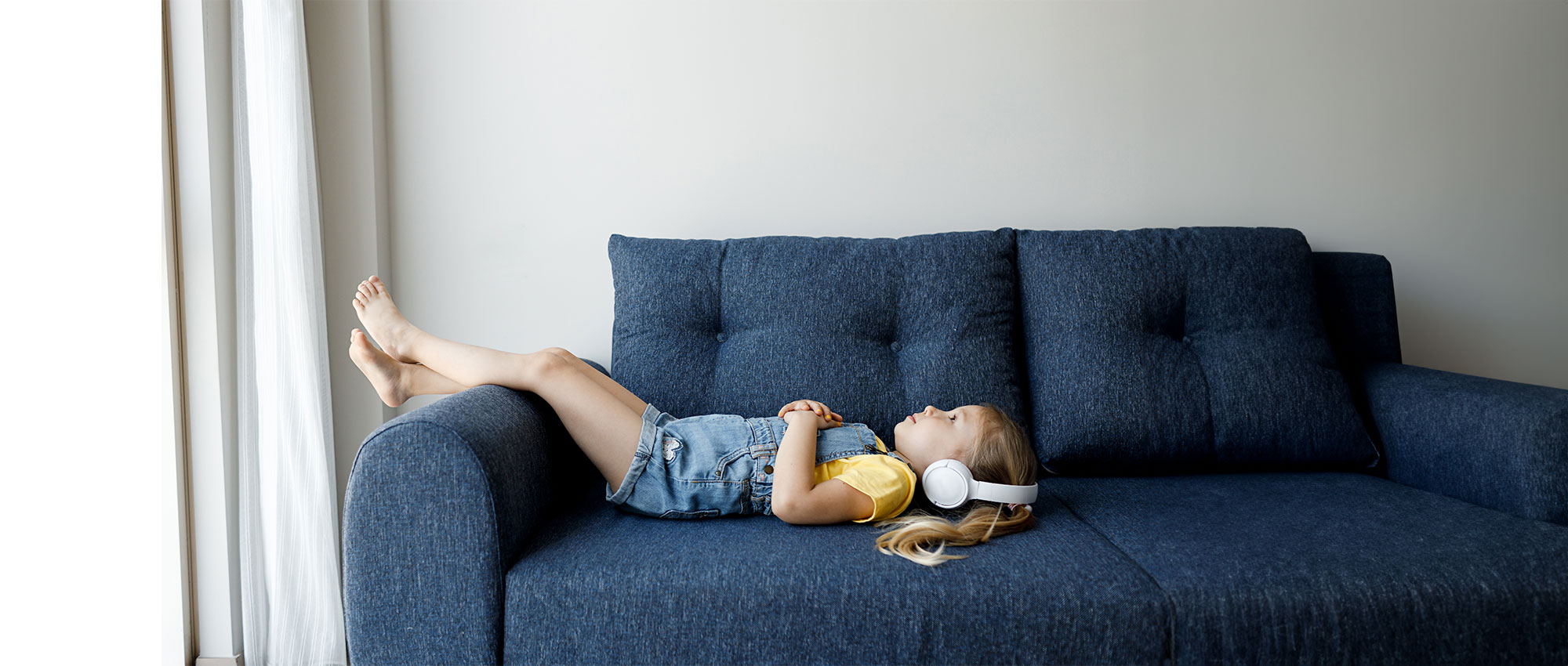 Ein Mädchen mit weißen Kopfhörern liegt entspannt auf einem blauen Sofa in hellem Raum und hört Musik.​​​​​​​​​​​​​​​​​​​​​​​​​​​​​​​​​​​​​​​​​​​