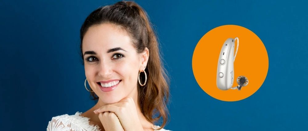 Junge Frau lächelt mit modernem Signia Hörgerät vor blauem Hintergrund und orangefarbener Grafik.​​​​​​​​​​​​​​​​​​​​​​​​​​​​​​​​​​​​​​​​​​​​​​​​