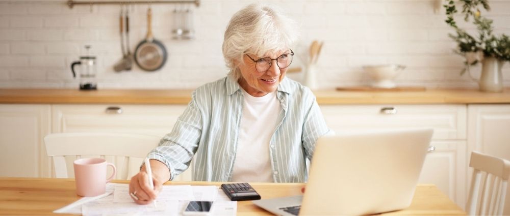 Ältere Frau sitzt in der Küche am Laptop und berechnet mit Taschenrechner steuerliche Vorteile bei Hörsystemerneuerung​​​​​​​​​​​​​​​​​​​​​​​​​​​