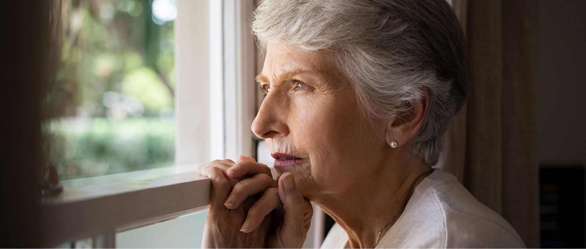 Ältere Frau mit grauen Haaren schaut nachdenklich aus dem Fenster in ruhiger Wohnumgebung, Symbol für soziale Isolation​​​​​​​​​​​​​​​​​​​​​​​​​​