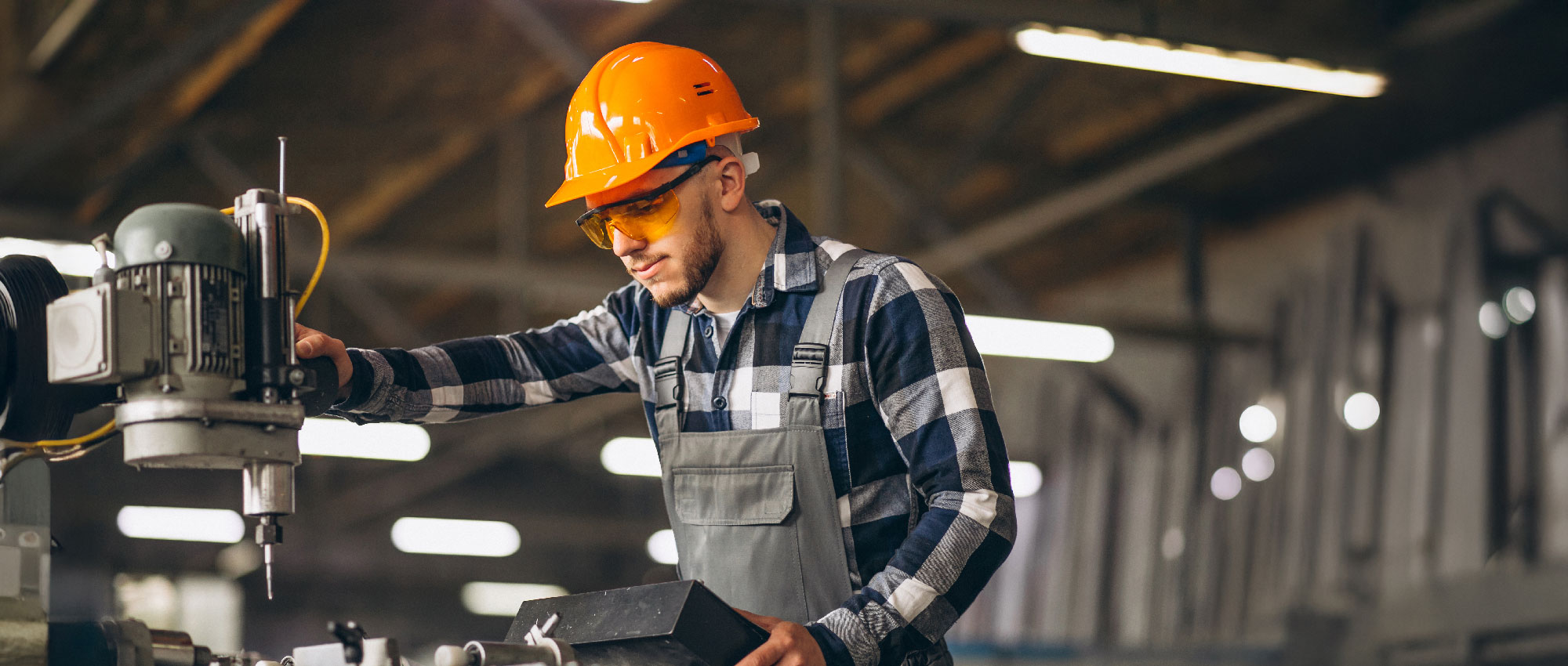 Handwerker mit Helm und Schutzbrille arbeitet an Industriemaschine in heller Werkstatt mit Holzdach.​​​​​​​​​​​​​​​​​​​​​​​​​​​​​​​​​​​​​​​​​​​​​