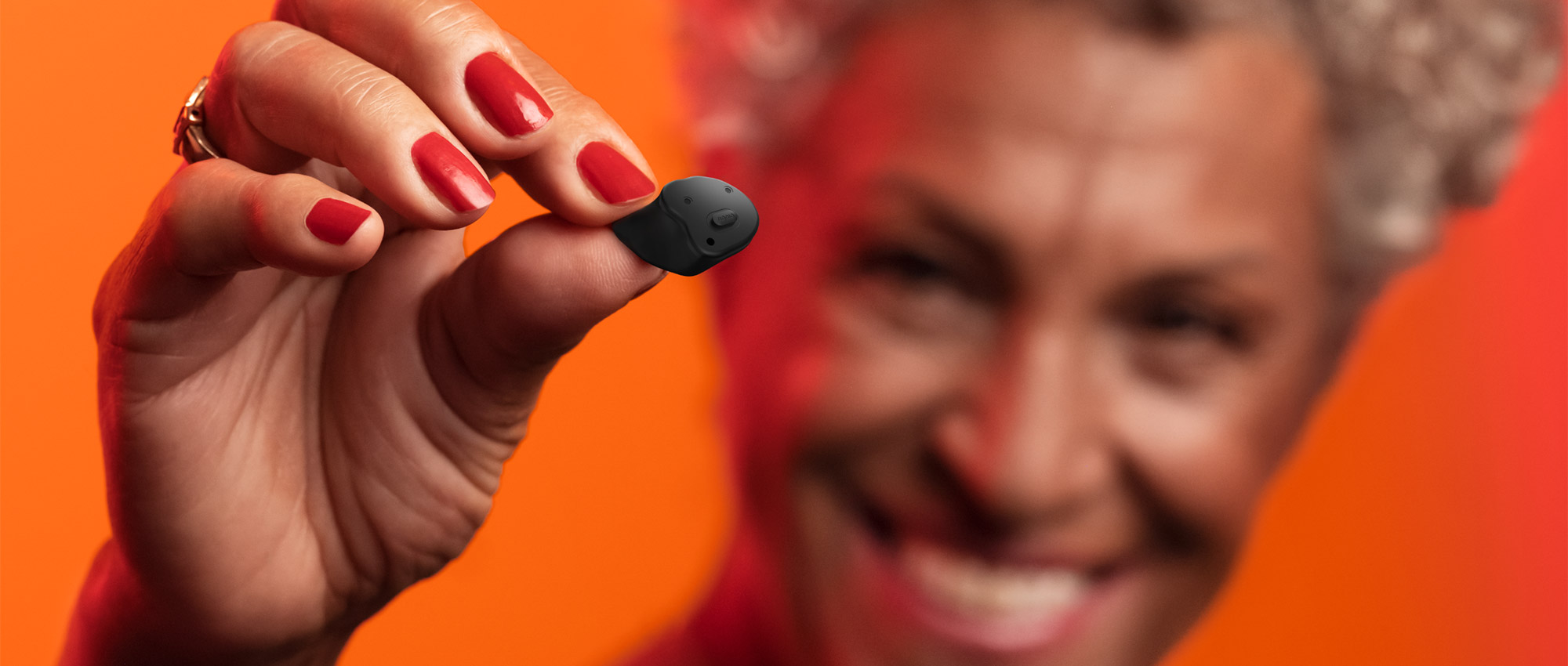 Nahaufnahme einer Hand mit roten Fingernägeln, die einen kleinen schwarzen Akku vor orangefarbenem Hintergrund hält​​​​​​​​​​​​​​​​​​​​​​​​​​​​​​