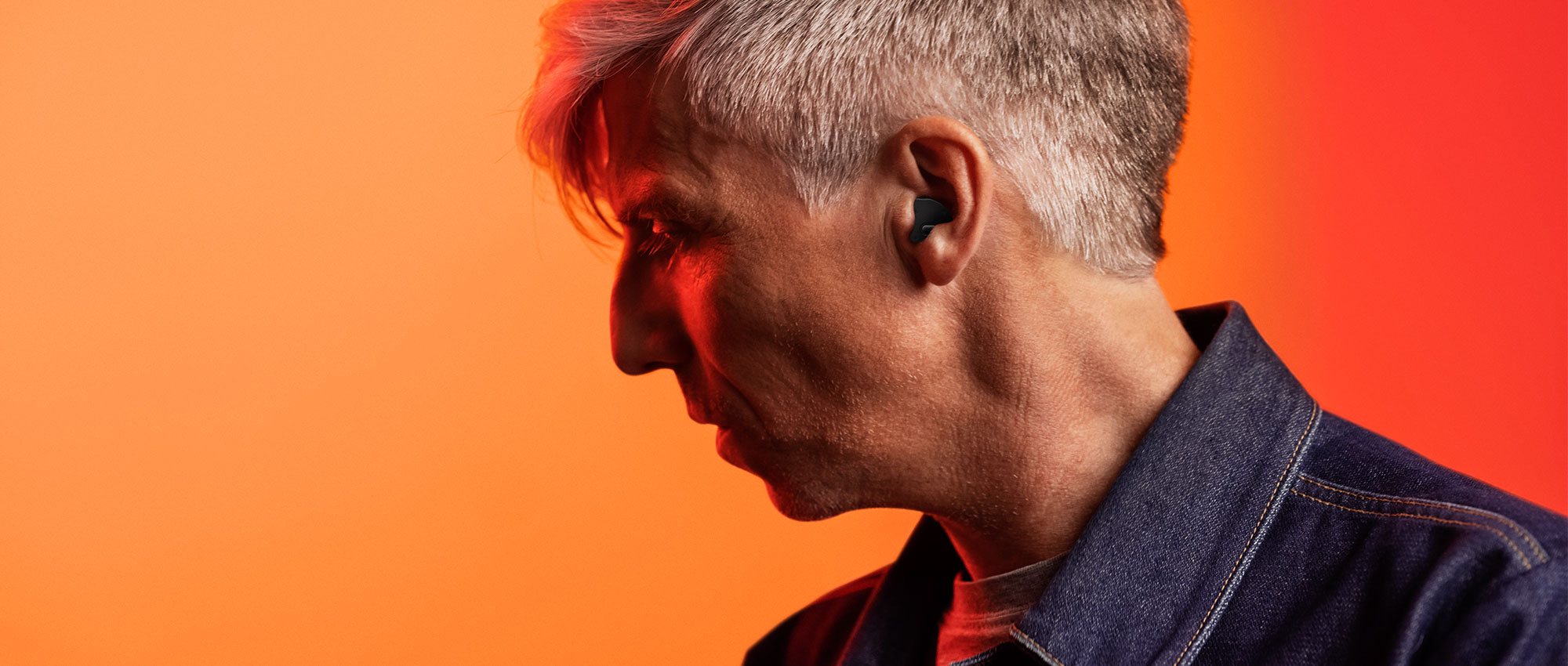 Seitenprofil eines Mannes mit grauem Haar und blauem Jeanshemd, der ein schwarzes Hörgerät trägt vor orange-rotem Hintergrund​​​​​​​​​​​​​​​​​​​​
