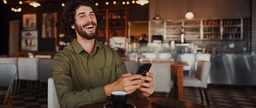Junger Mann sitzt in gemütlichem Café, lacht und hält Smartphone, vor ihm steht eine Kaffeetasse. Entspannte Atmosphäre​​​​​​​​​​​​​​​​​​​​​​​​​​