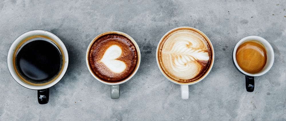 Vier Tassen mit schwarzem Kaffee, Cappuccino mit Herz, Latte Art und Espresso auf grauem Betonuntergrund​​​​​​​​​​​​​​​​​​​​​​​​​​​​​​​​​​​​​​​​​