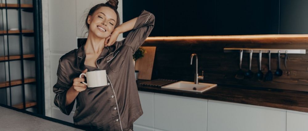 Junge Frau in dunklem Pyjama genießt Kaffee in heller Küche mit Holzarbeitsplatte und entspanntem Ausdruck​​​​​​​​​​​​​​​​​​​​​​​​​​​​​​​​​​​​​​​