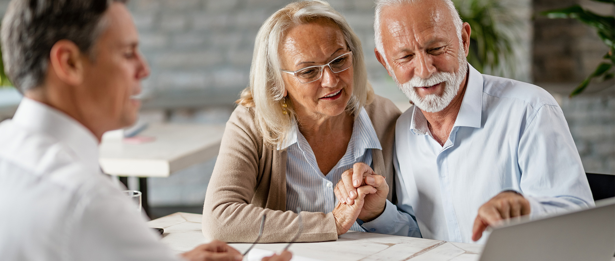 Älteres Paar sitzt mit Berater am Tisch und schaut gemeinsam auf Laptop. Beratungssituation in heller, freundlicher Umgebung.​​​​​​​​​​​​​​​​​​​​