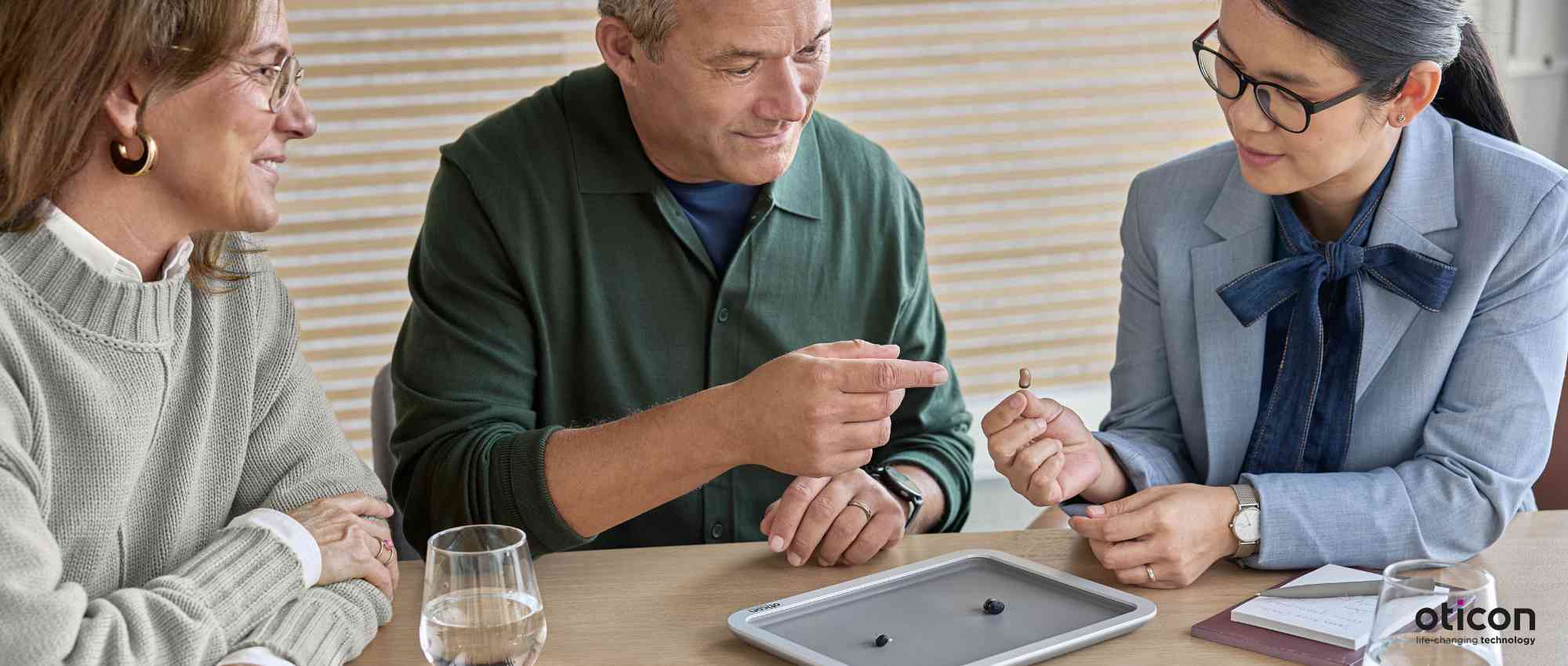 Drei Personen sitzen am Tisch und betrachten ein kleines Hörgerät, moderne Hörtechnologie im Gespräch​​​​​​​​​​​​​​​​​​​​​​​​​​​​​​​​​​​​​​​​​​​​