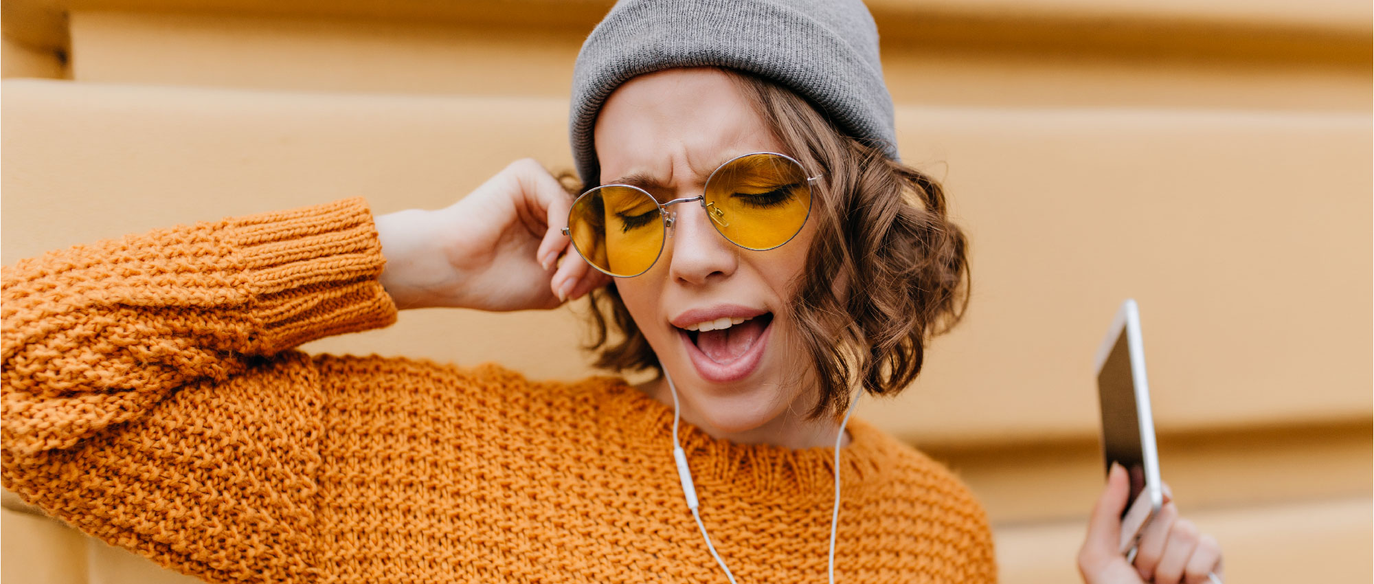 Junge Frau mit grauer Mütze und gelben Sonnenbrillen hört Musik über Kopfhörer und hält Smartphone in der Hand​​​​​​​​​​​​​​​​​​​​​​​​​​​​​​​​​​​