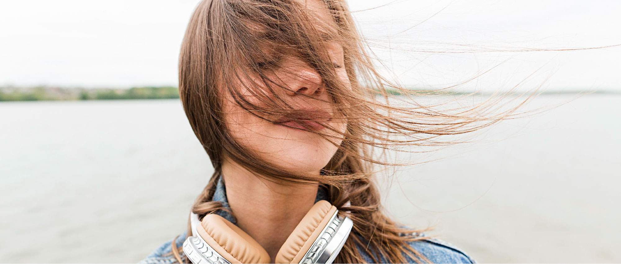 Junge Frau mit braunen Haaren und Kopfhörern steht am Seeufer, Haare wehen im Wind, entspannte Stimmung​​​​​​​​​​​​​​​​​​​​​​​​​​​​​​​​​​​​​​​​​​