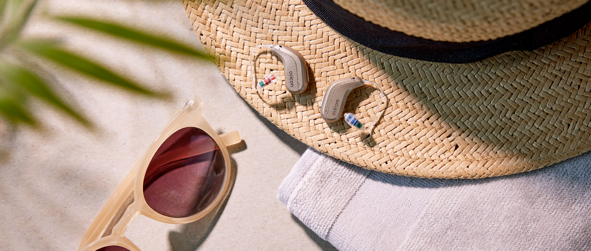 Zwei Oticon Hörgeräte liegen auf Strohhut neben Sonnenbrille und weißem Handtuch in sommerlicher Atmosphäre.​​​​​​​​​​​​​​​​​​​​​​​​​​​​​​​​​​​​​