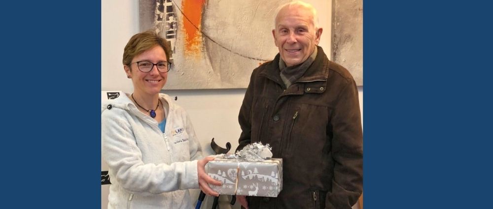 Frau übergibt älterem Mann ein Geschenkpaket vor abstraktem Bild in freundlicher Atmosphäre​​​​​​​​​​​​​​​​​​​​​​​​​​​​​​​​​​​​​​​​​​​​​​​​​​​​​​