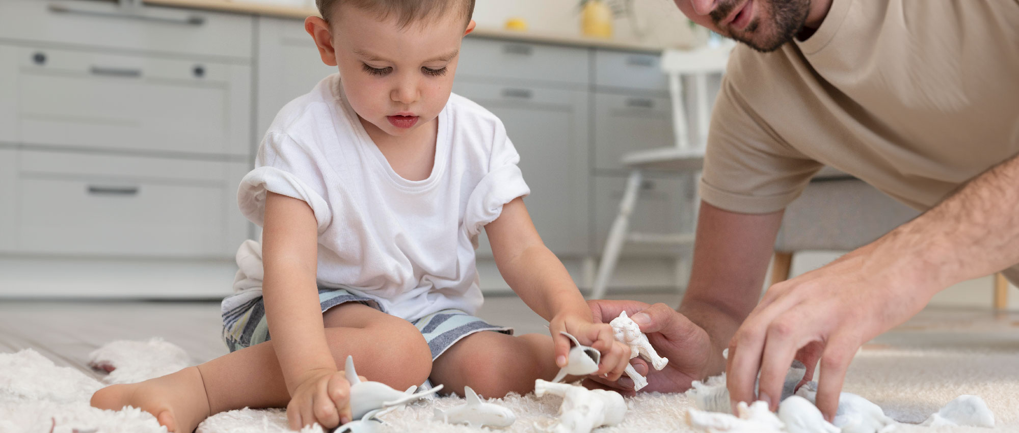 Kleiner Junge spielt mit weißen Tierfiguren auf Teppich in heller Küche, Vater unterstützt beim Geräuschspiel​​​​​​​​​​​​​​​​​​​​​​​​​​​​​​​​​​​​