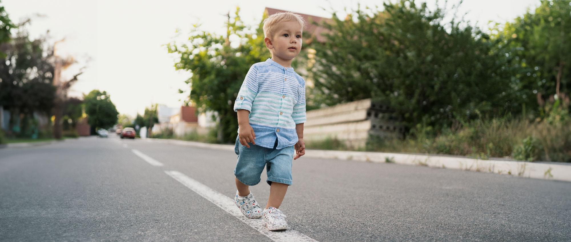 Kleiner Junge in blauem Hemd und Shorts läuft auf ruhiger Straße mit Häusern und Grün im Hintergrund​​​​​​​​​​​​​​​​​​​​​​​​​​​​​​​​​​​​​​​​​​​​​