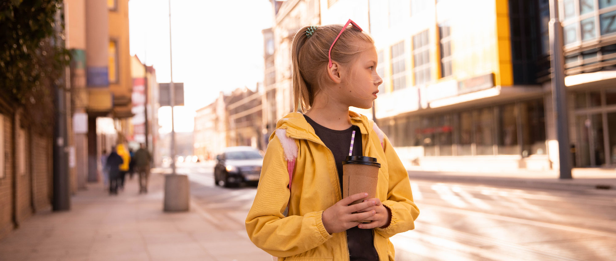 Schulkind mit gelber Jacke und Getränk steht auf sonnigem Bürgersteig in Stadt mit Gebäuden und Autos im Hintergrund​​​​​​​​​​​​​​​​​​​​​​​​​​​​​
