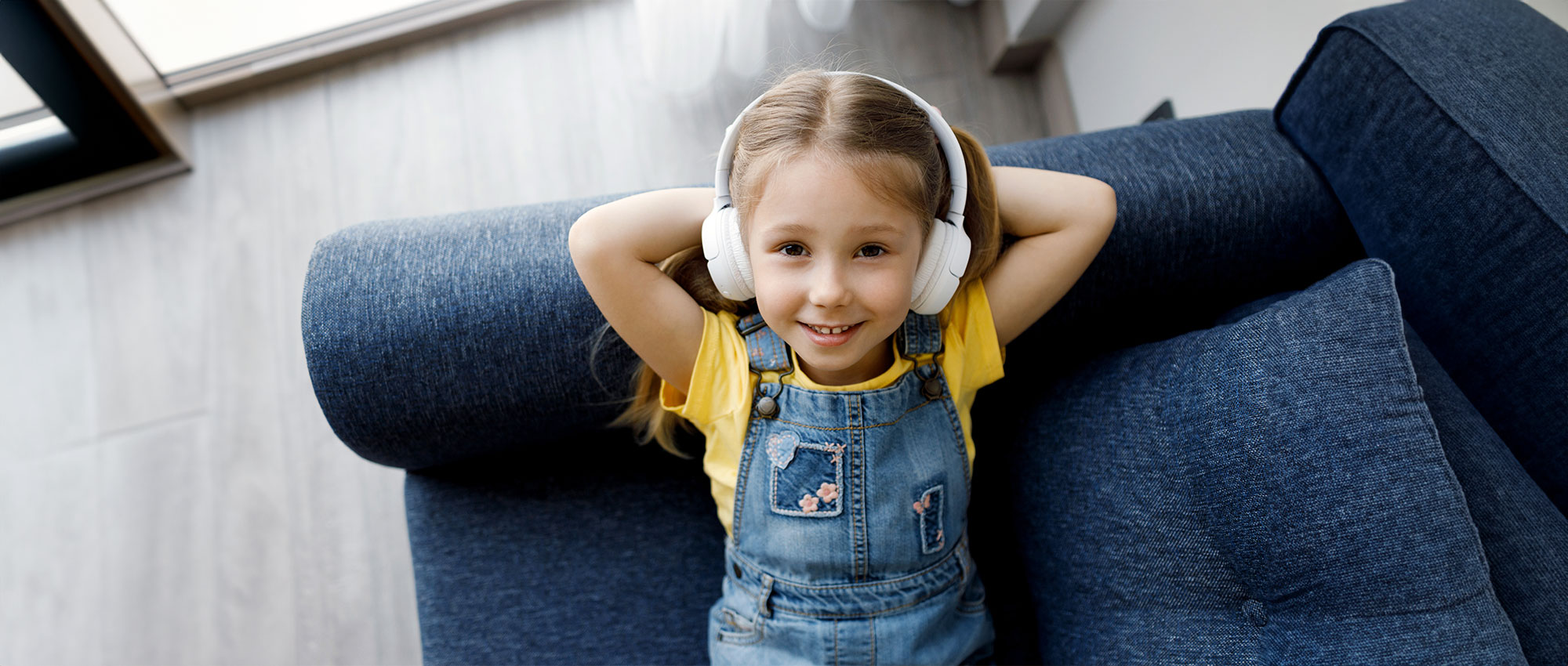 Ein Mädchen mit gelbem T-Shirt und Jeans-Latzhose entspannt auf blauem Sofa und hört Musik mit weißen Kopfhörern​​​​​​​​​​​​​​​​​​​​​​​​​​​​​​​​​