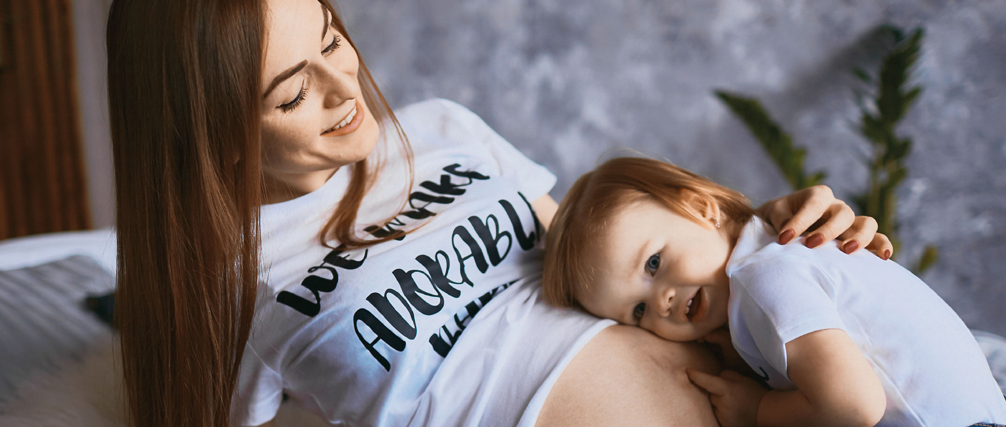 Schwangere Frau liegt lächelnd mit kleinem Kind auf dem Bauch in gemütlicher Wohnumgebung mit Pflanze im Hintergrund​​​​​​​​​​​​​​​​​​​​​​​​​​​​​