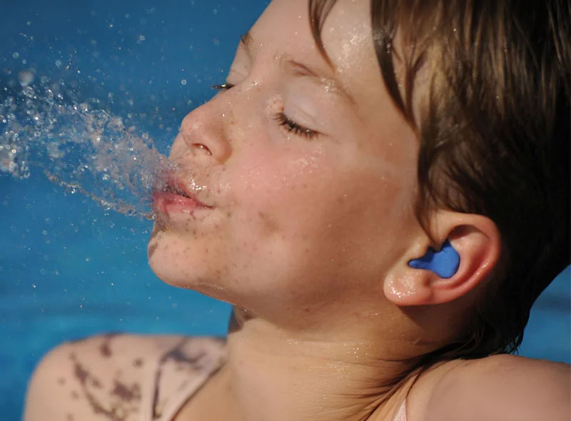 Kind mit blauem Schwimmschutz und Gehörschutz spritzt Wasser im Schwimmbad, Sommer und Wasserspaß im Fokus.​​​​​​​​​​​​​​​​​​​​​​​​​​​​​​​​​​​​​​