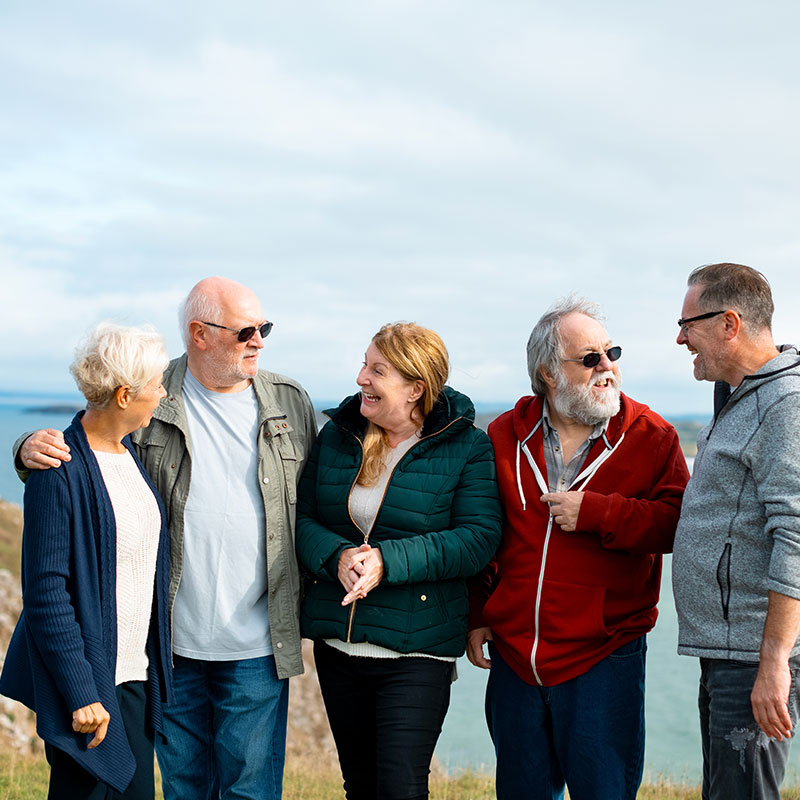 Fünf ältere Personen unterhalten sich lachend an der Küste bei bewölktem Himmel. Gruppenstimmung und Naturkulisse sichtbar.​​​​​​​​​​​​​​​​​​​​​​