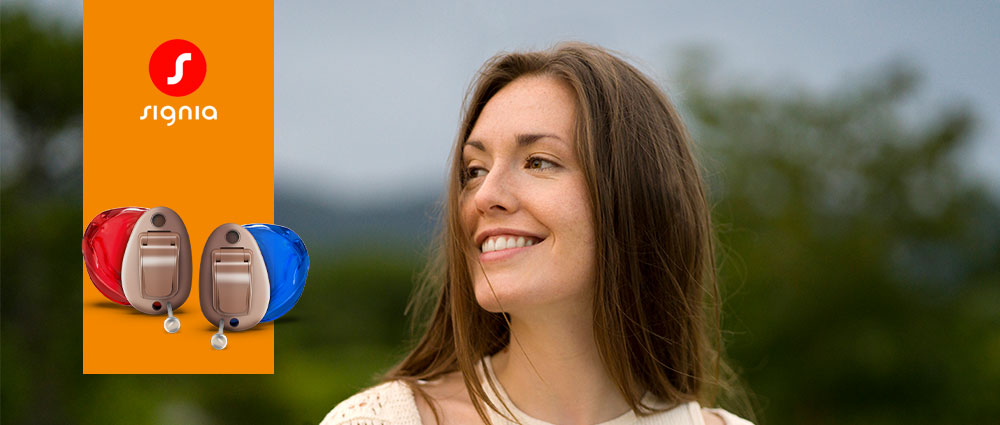 Frau lächelt im Freien neben Signia Insio Charge Go CIC IX Hörgeräten in Rot und Blau vor grünem Hintergrund​​​​​​​​​​​​​​​​​​​​​​​​​​​​​​​​​​​​​