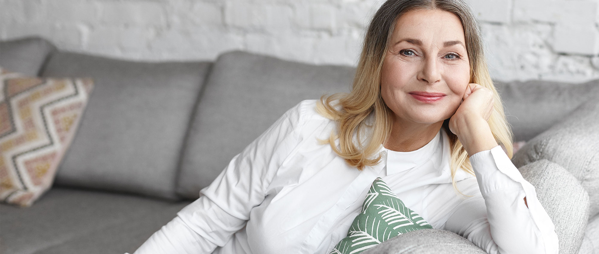 Ältere Frau sitzt entspannt auf grauem Sofa mit gemusterten Kissen in hellem Wohnzimmer. Ruhige Atmosphäre​​​​​​​​​​​​​​​​​​​​​​​​​​​​​​​​​​​​​​​