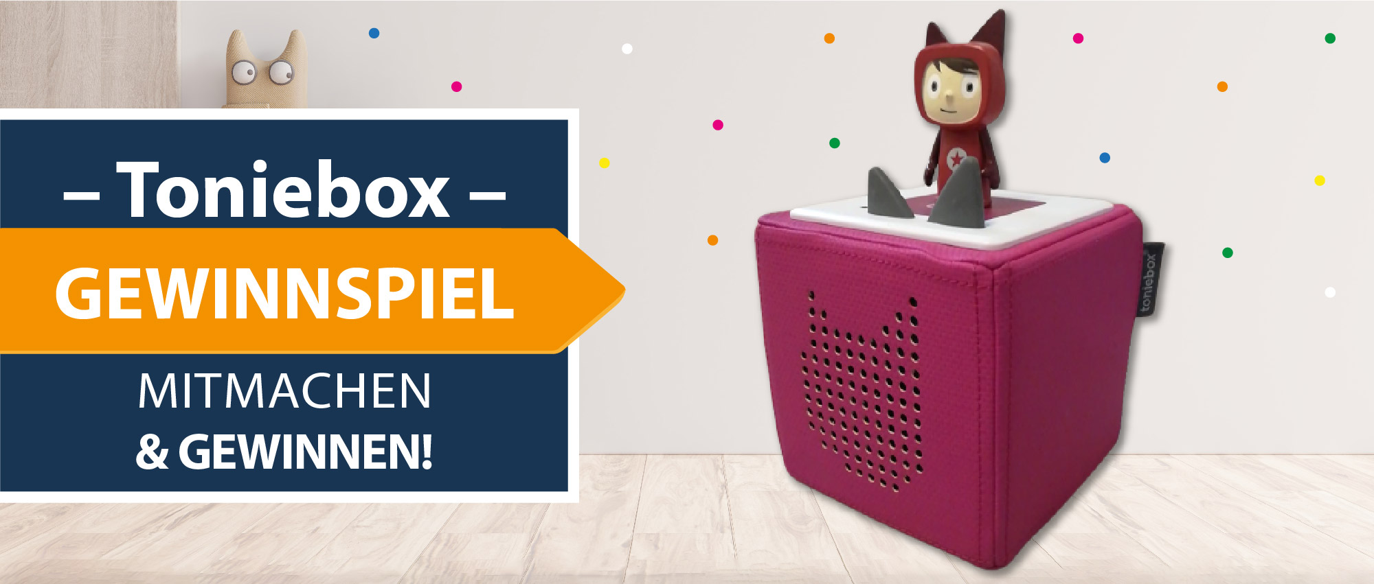 Rosa Toniebox mit roter Spielfigur auf hellem Holzfußboden vor weißer Wand mit bunten Punkten.​​​​​​​​​​​​​​​​​​​​​​​​​​​​​​​​​​​​​​​​​​​​​​​​​​​