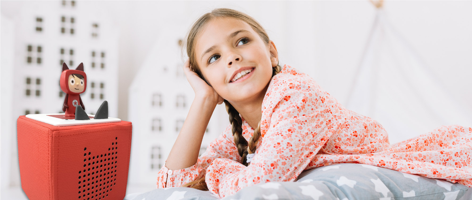 Ein Mädchen liegt entspannt neben einer roten Toniebox mit Spielfigur in einem hellen, kindgerechten Zimmer.​​​​​​​​​​​​​​​​​​​​​​​​​​​​​​​​​​​​​