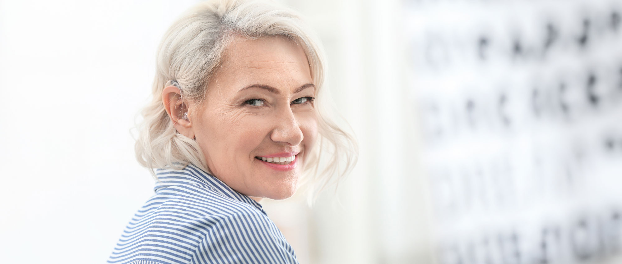 Ältere Frau mit weißem Haar trägt ein Hörgerät und lächelt freundlich vor hellem Hintergrund mit Sehtesttafel.​​​​​​​​​​​​​​​​​​​​​​​​​​​​​​​​​​​