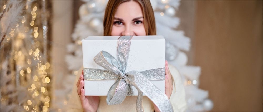 Junge Frau zeigt Geschenk mit silberner Schleife vor festlich geschmücktem Weihnachtsbaum.​​​​​​​​​​​​​​​​​​​​​​​​​​​​​​​​​​​​​​​​​​​​​​​​​​​​​​​