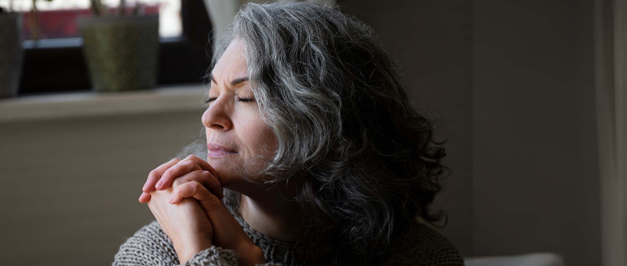 Ältere Frau mit grauen Haaren sitzt entspannt mit geschlossenen Augen und gefalteten Händen in einem gemütlichen Raum​​​​​​​​​​​​​​​​​​​​​​​​​​​​