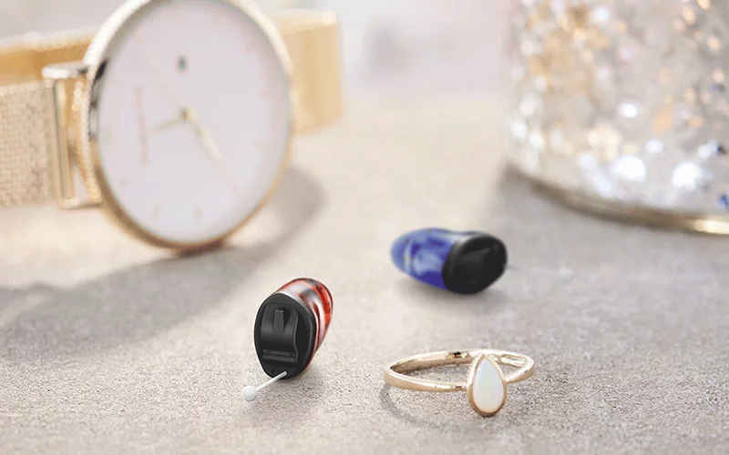 Zwei kleine In-Ear-Hörgeräte neben einer Uhr und einem Ring. Diskrete Hörlösungen, In-Ear-Hörgeräte, elegantes Hörgerätedesign.