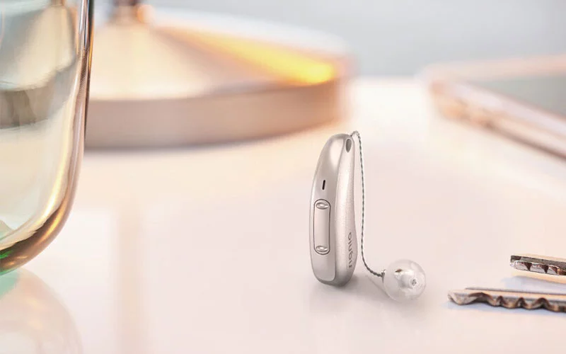 Ein Signia-Hörgerät steht auf einer Oberfläche neben einem Glas und Schlüsseln. Signia-Hörgeräte, fortschrittliche Hörtechnologie, elegantes Design.