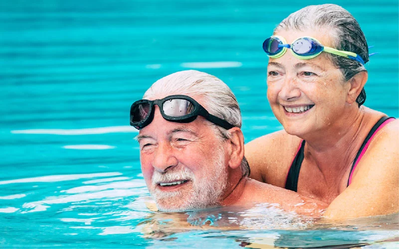 Älteres Paar genießt das Schwimmen in einem Pool, beide lächeln und tragen Schwimmbrillen. Hörgeräte für Senioren, aktives Leben mit Hörgeräten, Schwimmen und Hören.