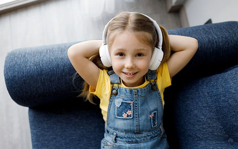 Kleines Mädchen entspannt sich mit Kopfhörern auf einem Sofa. Kinderhörgeräte, Hörkomfort zu Hause, kindgerechte Hörlösungen.