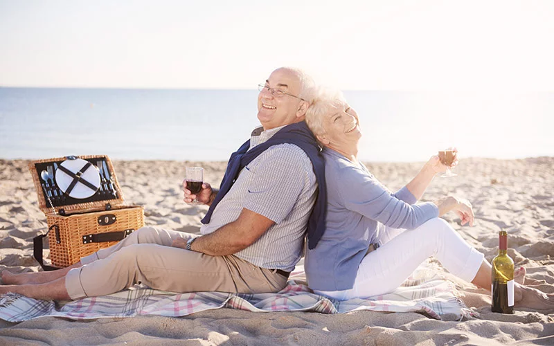 Älteres Paar genießt ein Picknick am Strand, beide lächelnd und entspannt. Hörkomfort im Freien, Hörgeräte für den Urlaub.