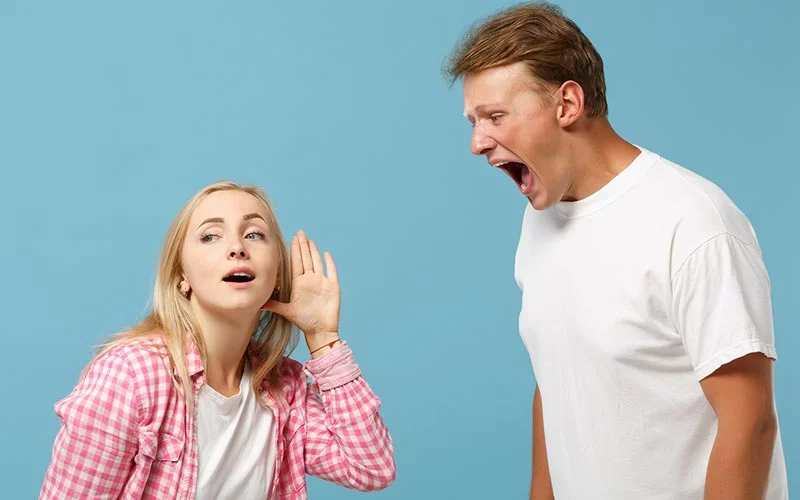 Frau hält die Hand ans Ohr, während ein Mann laut spricht. Hörprobleme, Notwendigkeit von Hörgeräten erkennen.